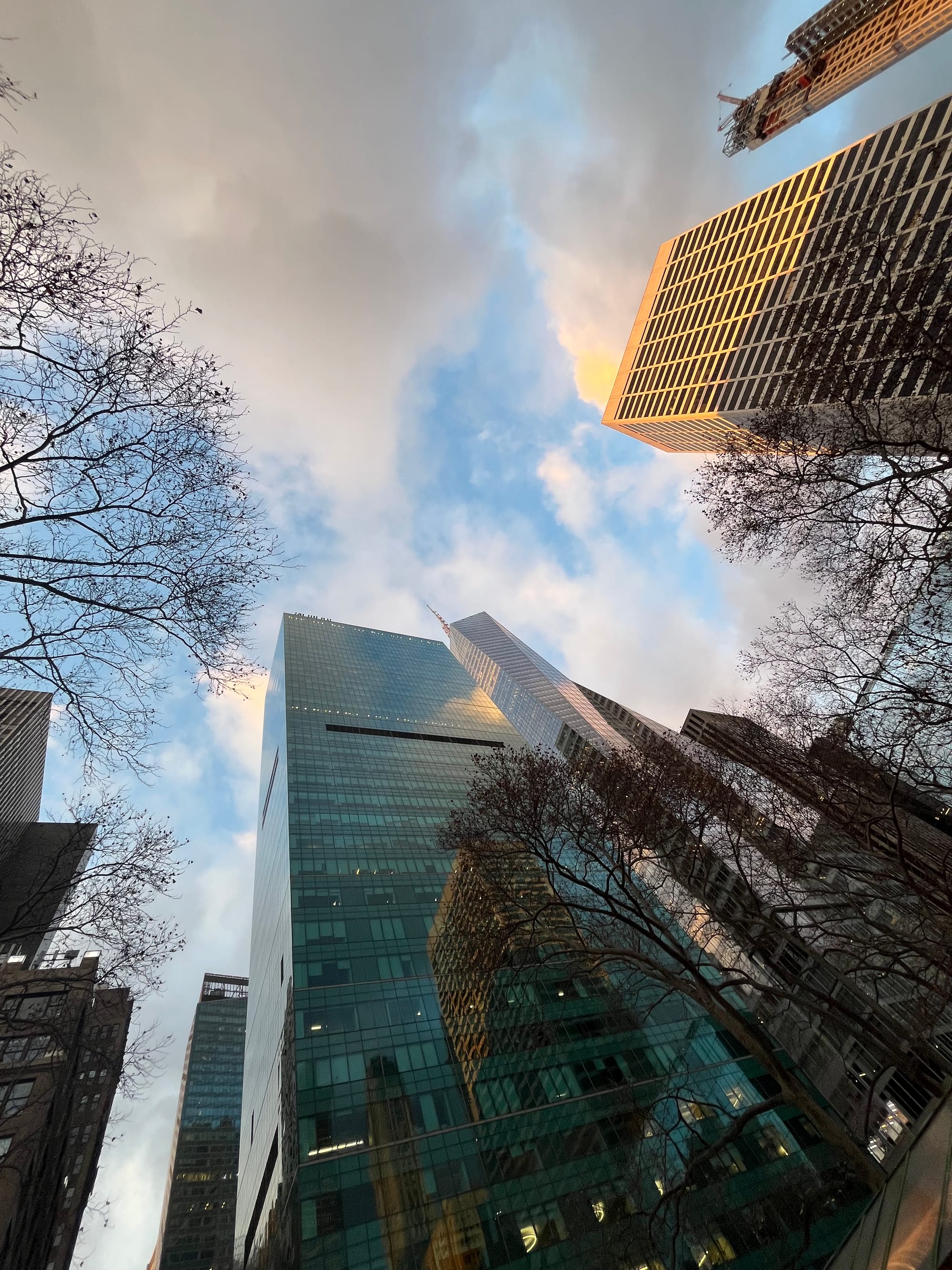 A cluster of glass skyscrapers reflecting other tall buildings. The sky is overcast with clouds, with the last rays of the evening sun shining down on them. 